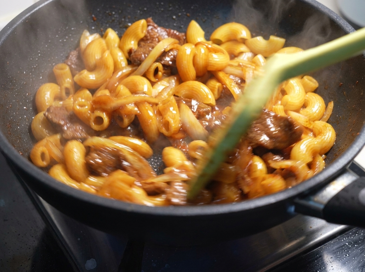 Macarrones, ternera y cebollas siendo salteados rápidamente con una espátula en una sartén caliente.