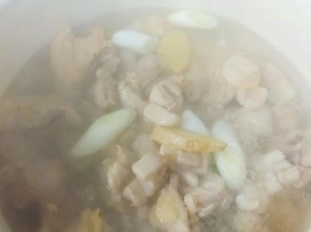 Agua caliente humeante en una olla blanca sobre trozos de pollo, jengibre y cebolletas.