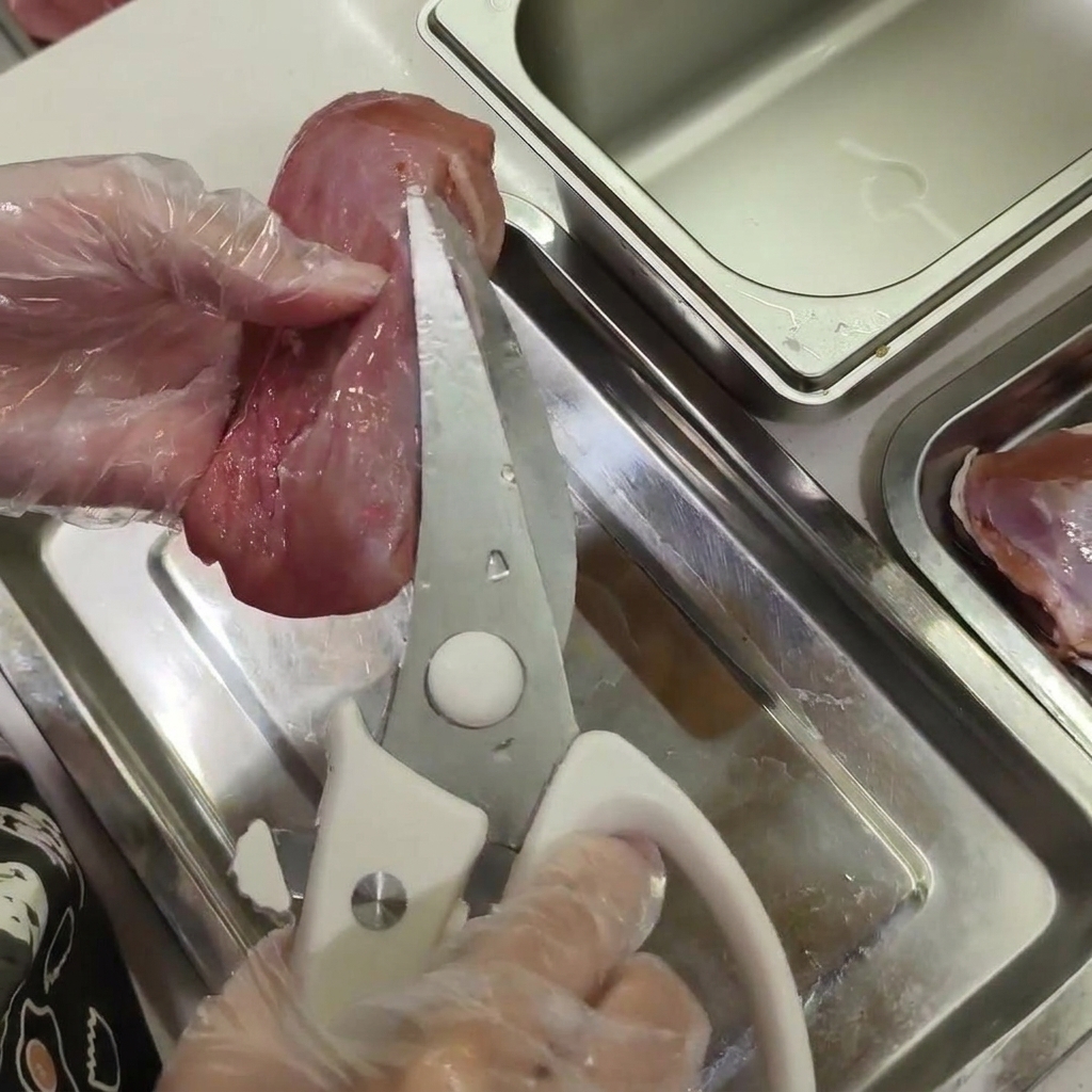 Manos con guantes sosteniendo carne de pato cruda mientras usan tijeras de cocina para recortar trozos pequeños de grasa blanca.