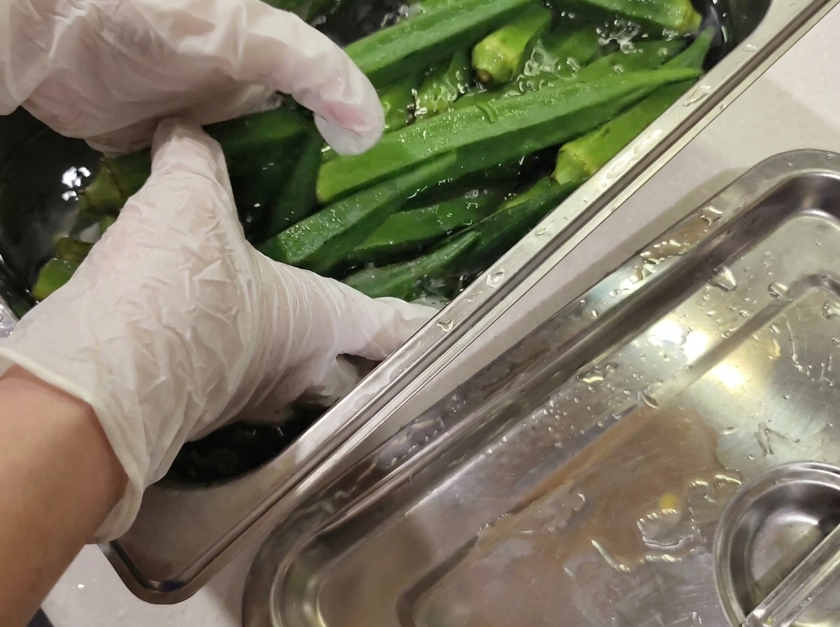 Manos con guantes lavando suavemente vainas de okra verde fresca en un recipiente de agua de acero inoxidable.