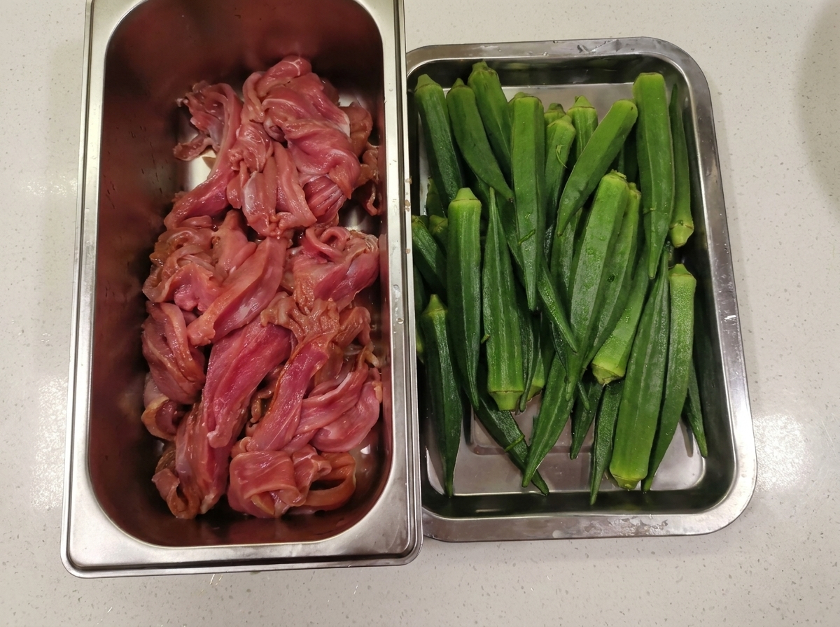 Un recipiente de metal con tiras de carne de pato junto a una bandeja llena de okra verde lavada.