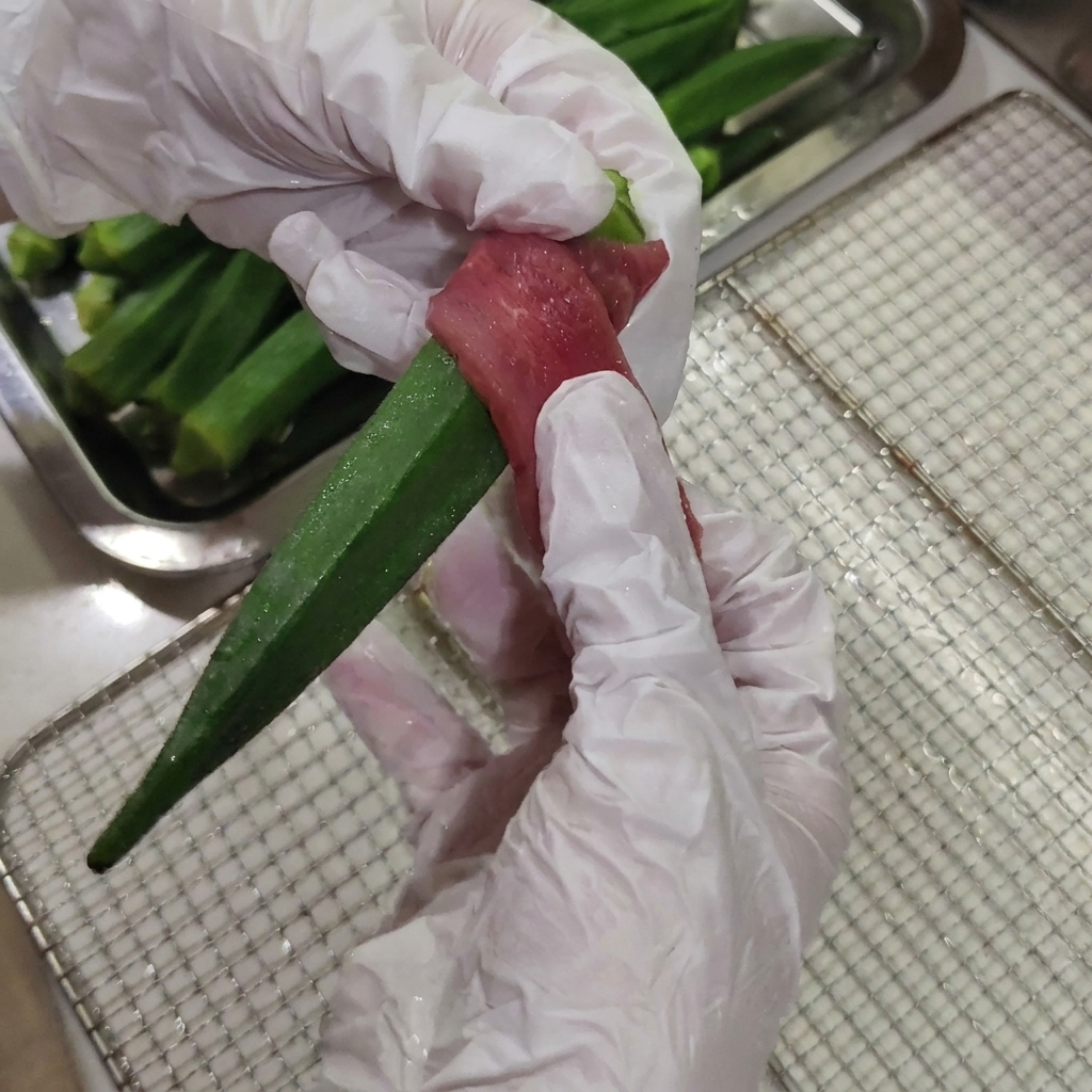 Manos con guantes envolviendo firmemente una tira fina de carne de pato cruda alrededor del centro de una vaina de okra verde.
