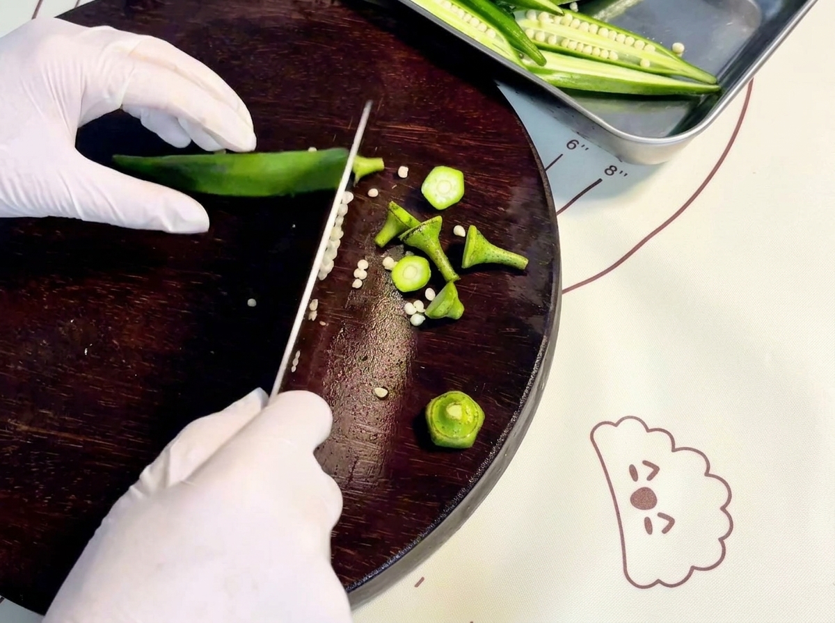 Manos con guantes cortando okra verde fresca a lo largo sobre una tabla de cortar de madera oscura.cite: 1