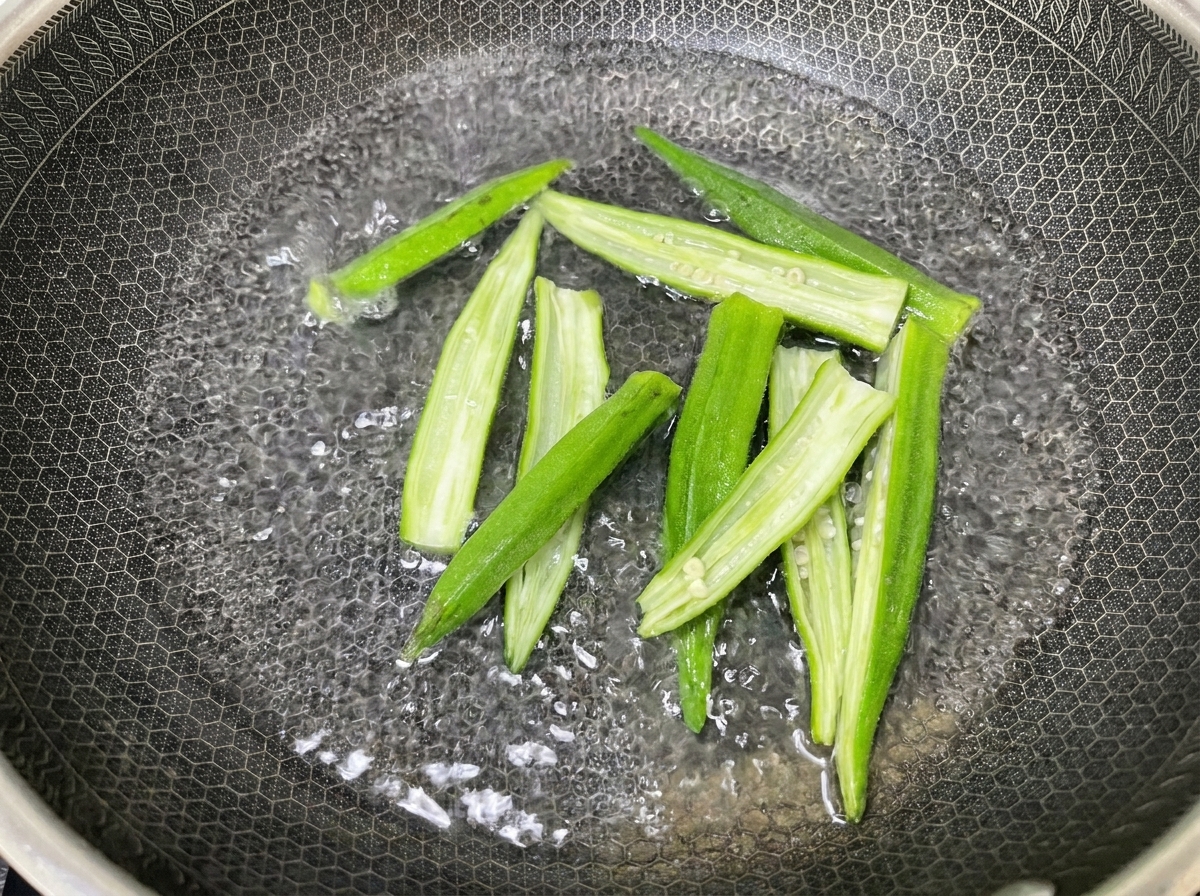 Mitades de okra huecas siendo blanqueadas en una olla de metal texturizado llena de agua hirviendo.cite: 1