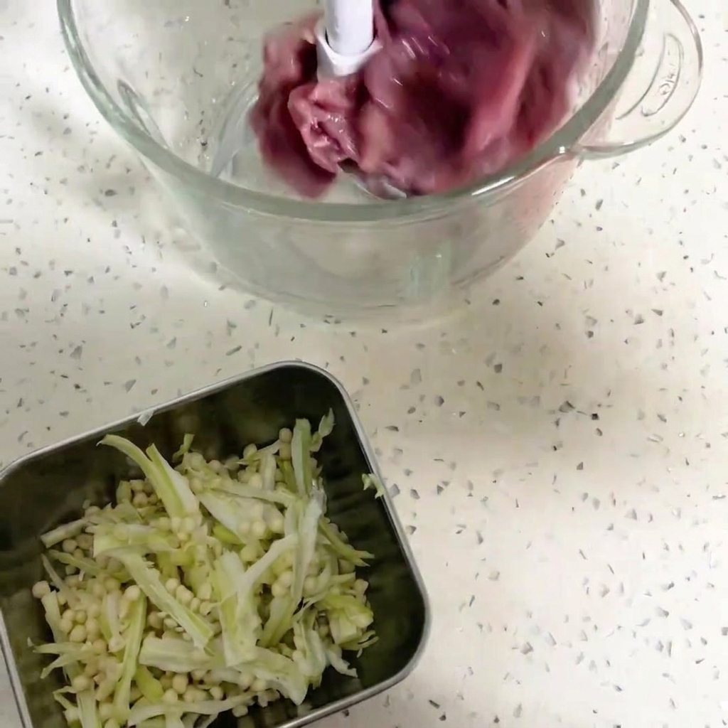 Un primer plano del recipiente de un procesador de alimentos que contiene trozos de carne de pato roja oscura y semillas de okra, listos para ser triturados en una pasta fina.cite: 1