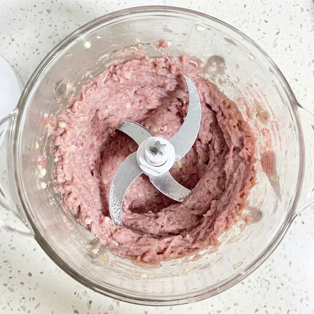 Una pasta de carne de pato cruda, de color rosa claro, finamente triturada y suave, dentro del recipiente de cristal transparente de un procesador de alimentos.cite: 1