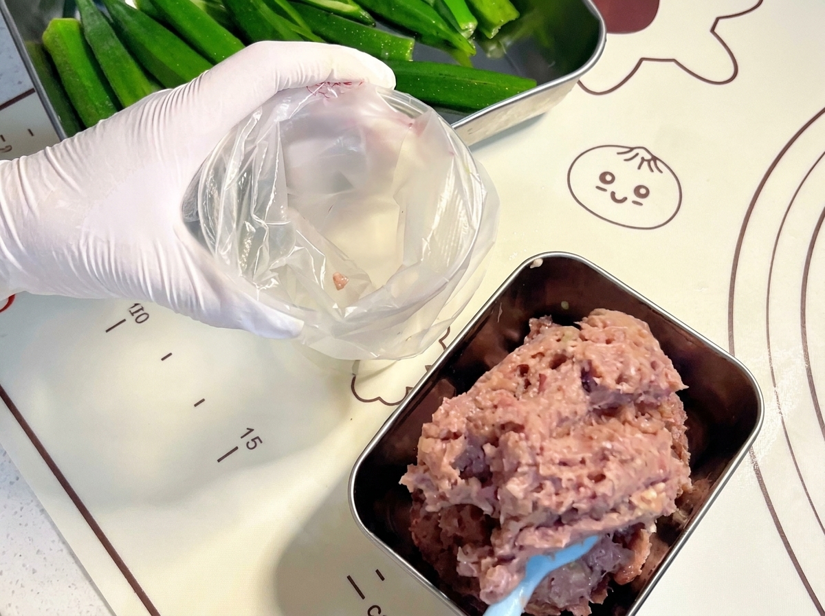 Manos con guantes usando una espátula azul para transferir la pasta de carne cruda triturada a una manga pastelera de plástico transparente.cite: 1