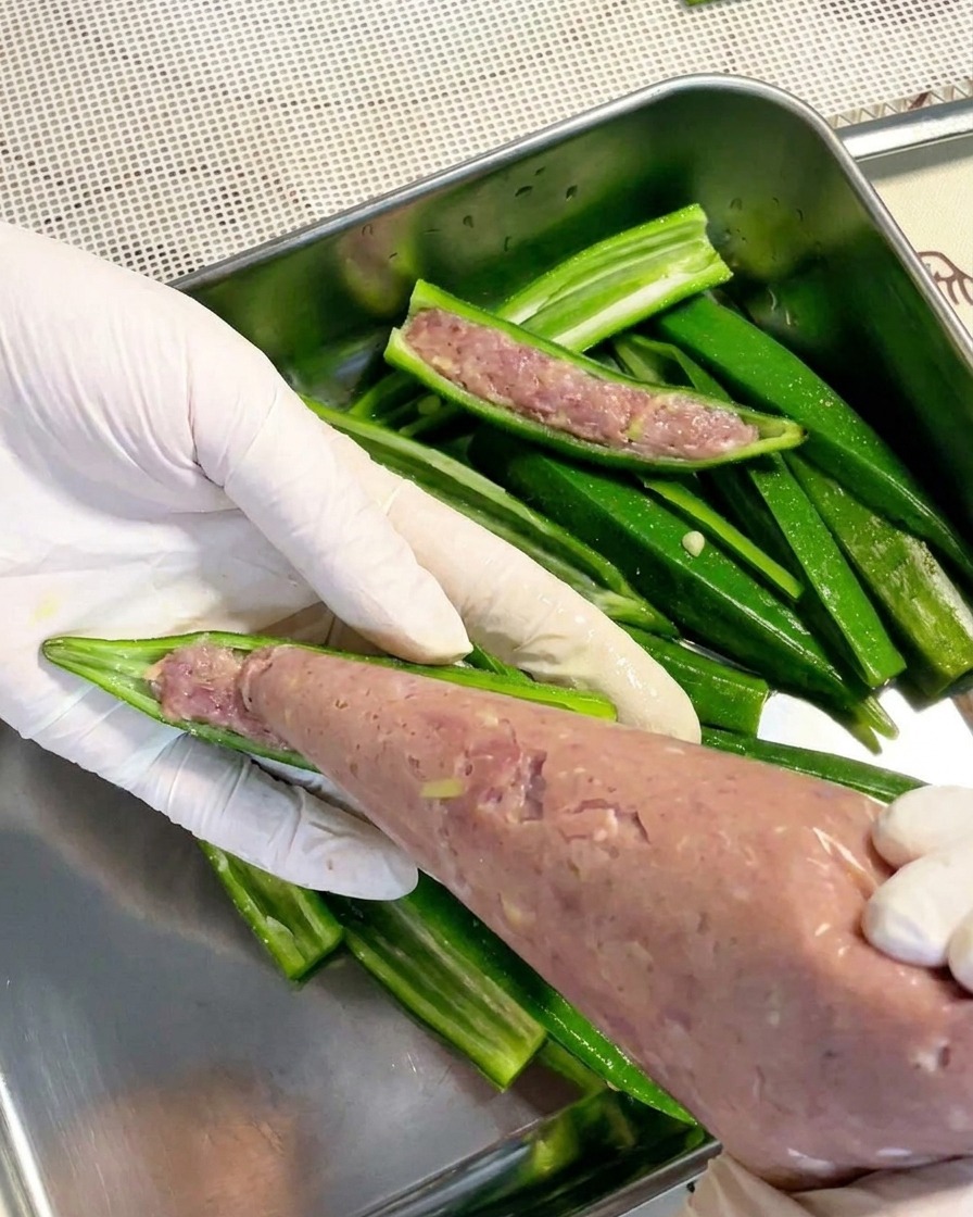 Un par de manos con guantes utilizando una manga pastelera transparente para rellenar una mitad de okra blanqueada con una pasta de carne suave y de color claro.cite: 1