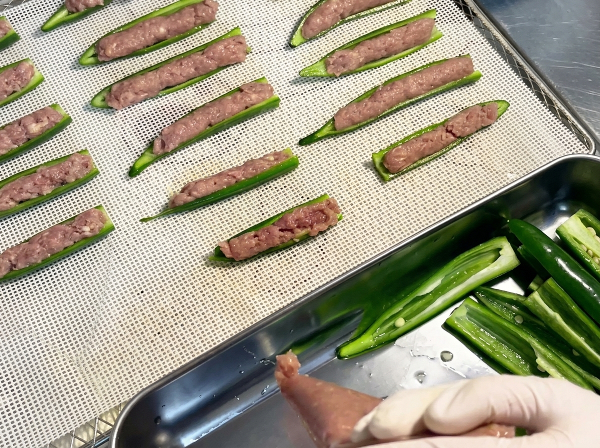 Barquitas de okra crudas rellenas, dispuestas ordenadamente en filas sobre la bandeja de malla de plástico blanco de un deshidratador.cite: 1