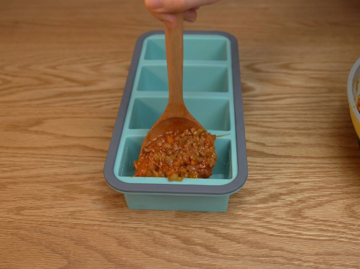 Salsa espesa de tomate y carne picada siendo servida en los compartimentos individuales de una bandeja de silicona azul.