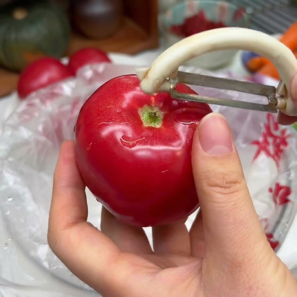 Manos sosteniendo un tomate rojo vibrante mientras usan un pelador de verduras blanco para quitar la piel.