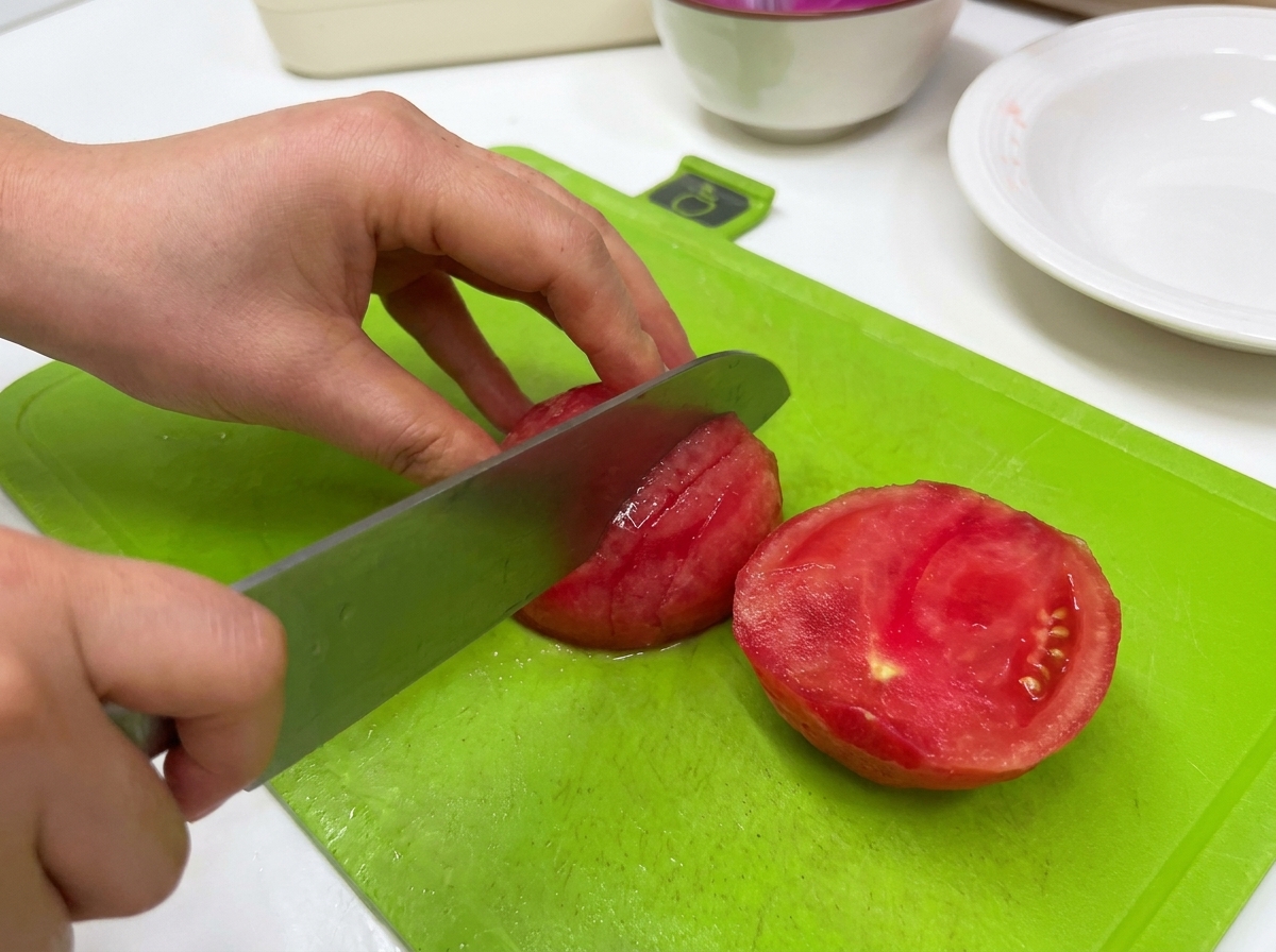 Una persona usando un cuchillo de cocina grande para rebanar un tomate pelado sobre una tabla de cortar verde.