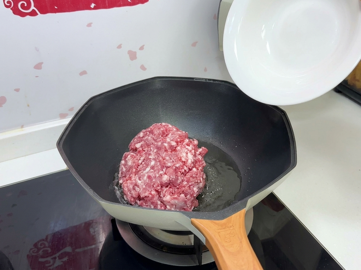 Un montón de carne de cerdo picada cruda cayendo desde un cuenco blanco hacia una sartén caliente con aceite.