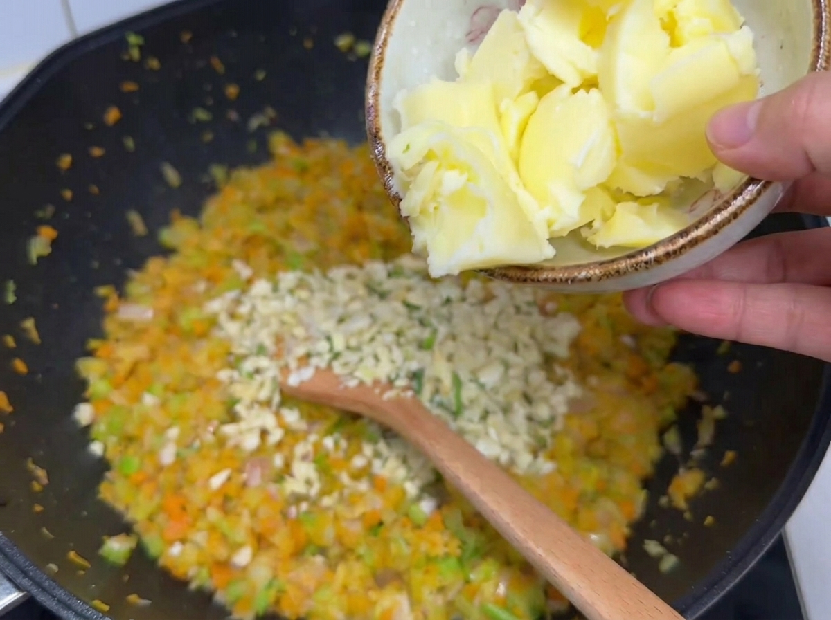 Trozos grandes de mantequilla vertidos desde un cuenco pequeño sobre las verduras picadas ablandadas y el ajo picado en un wok.