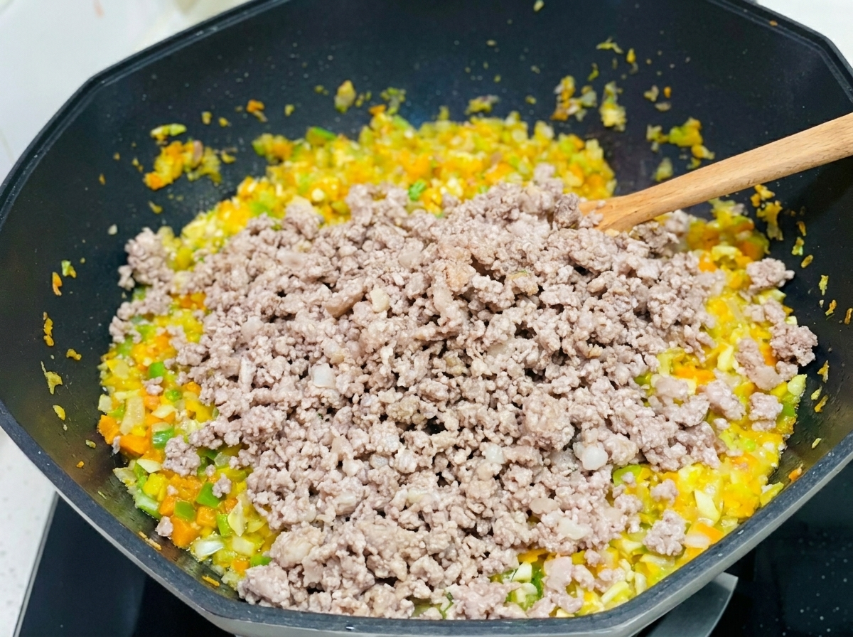 Carne de cerdo picada siendo incorporada a una mezcla de zanahorias, apio y cebollas salteadas en un wok grande.