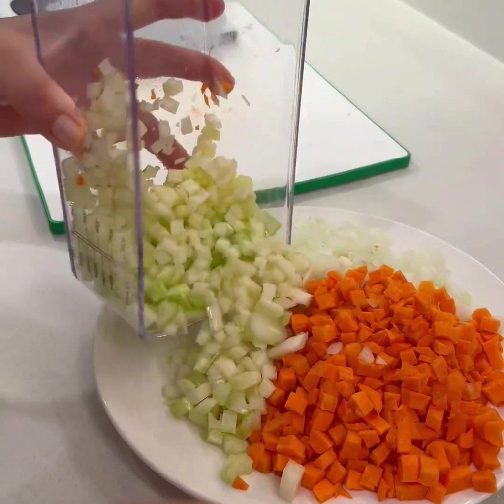 Apio picado vaciado de un picador transparente a un plato junto a zanahorias y cebollas picadas.