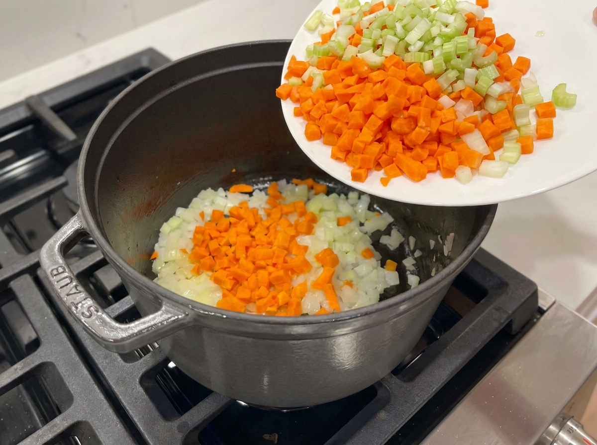 Zanahorias y apio picados vertidos desde un plato blanco a una olla con cebollas salteadas.