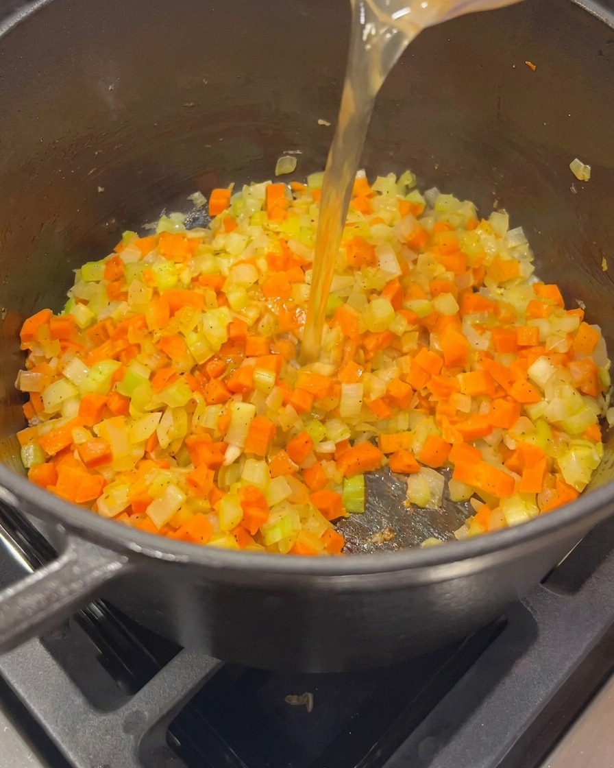 Caldo de pollo dorado vertiéndose desde una taza medidora de cristal a una olla que contiene cebollas, zanahorias y apio salteados.