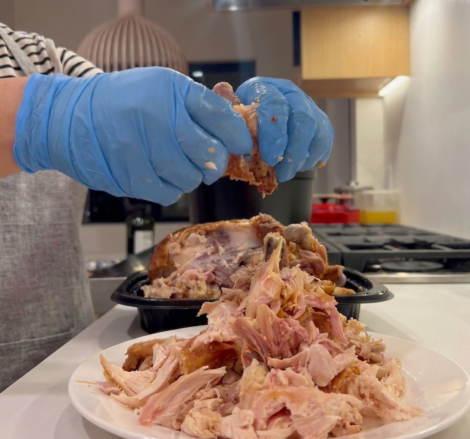 Manos con guantes culinarios azules desmenuzando carne de pollo asado cocido en un montón sobre un plato blanco.