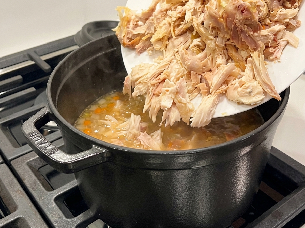 Pollo desmenuzado añadiéndose desde un plato blanco a una olla negra de sopa hirviendo a fuego lento.