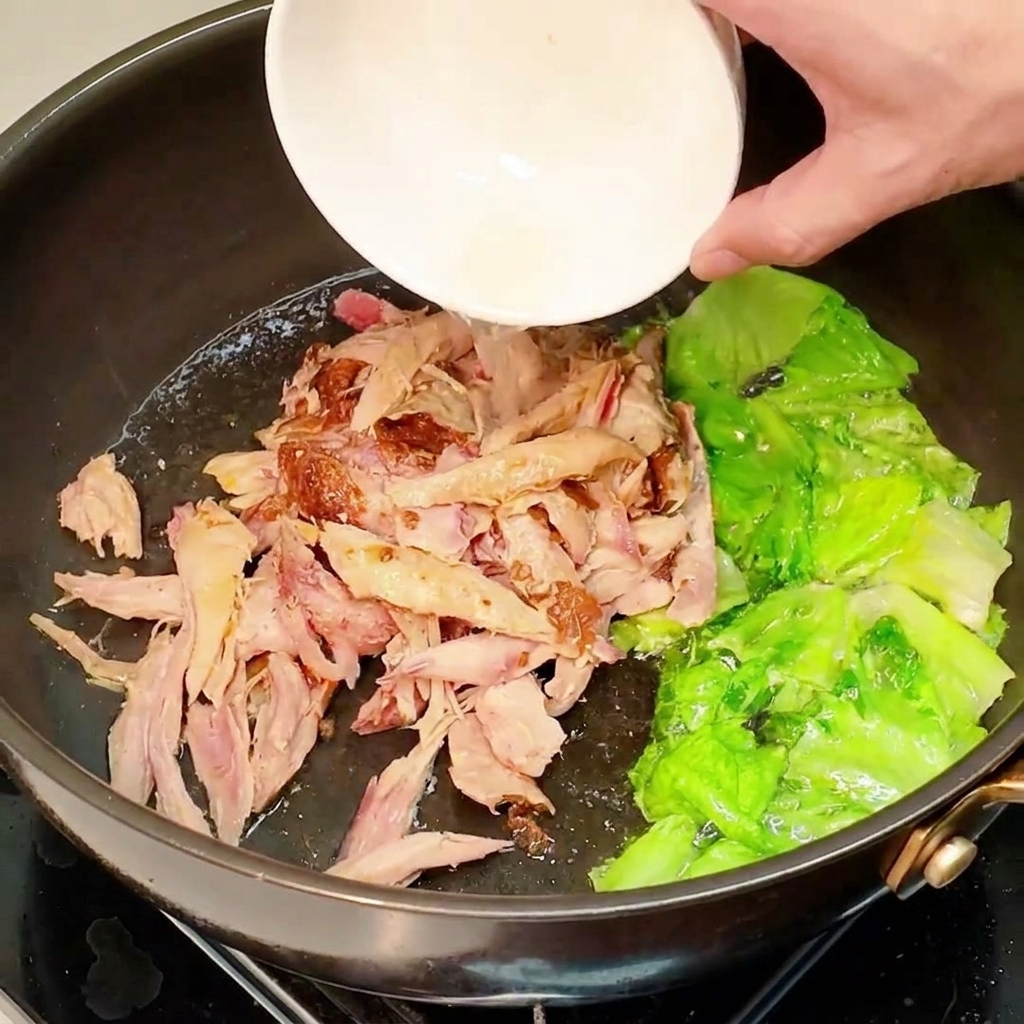 Pollo a la pimienta desmenuzado y precocido vertiéndose desde un pequeño bol blanco a una sartén con lechuga salteada.