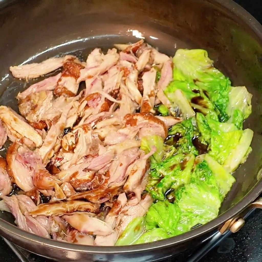 Pollo desmenuzado y lechuga cubiertos de salsa de soja oscura mezclándose en una sartén.