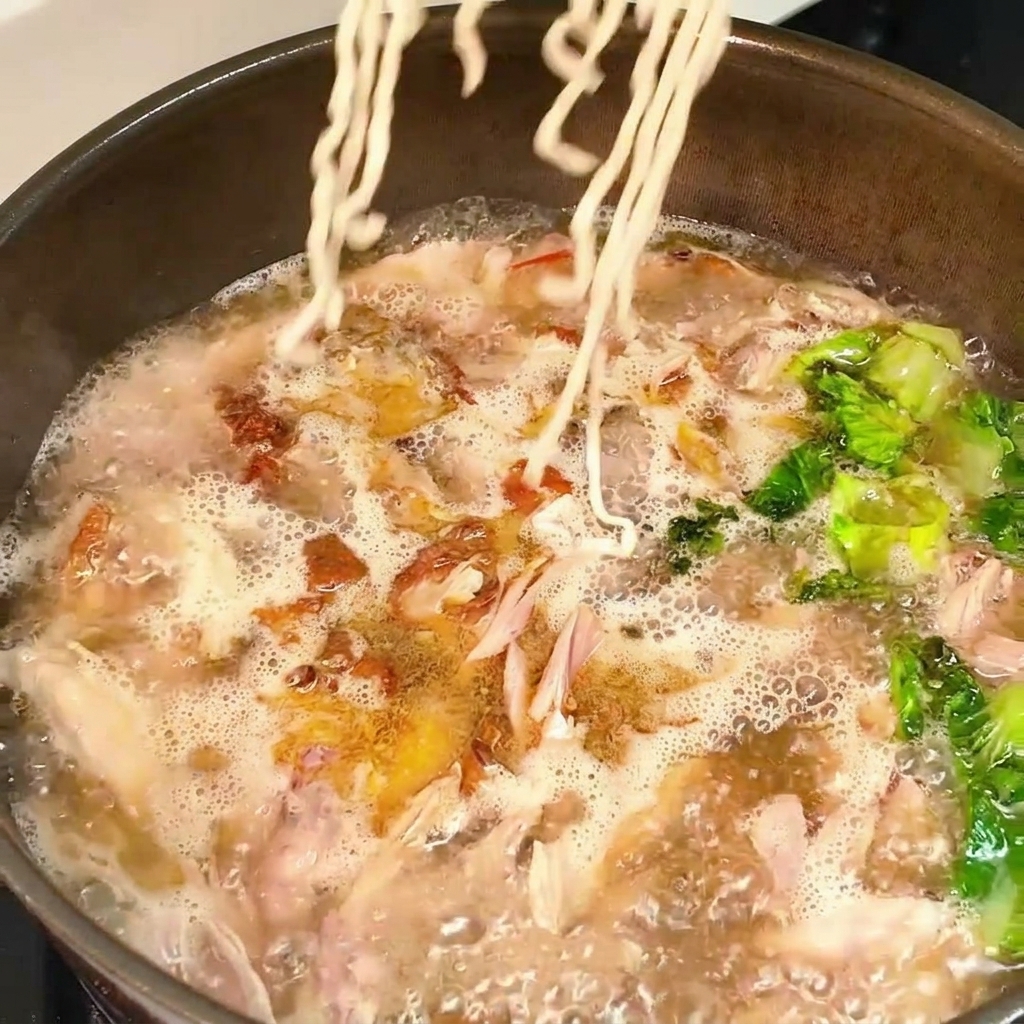 Un bloque de fideos secos ondulados introduciéndose en una sartén con caldo de pollo y lechuga hirviendo a borbotones.