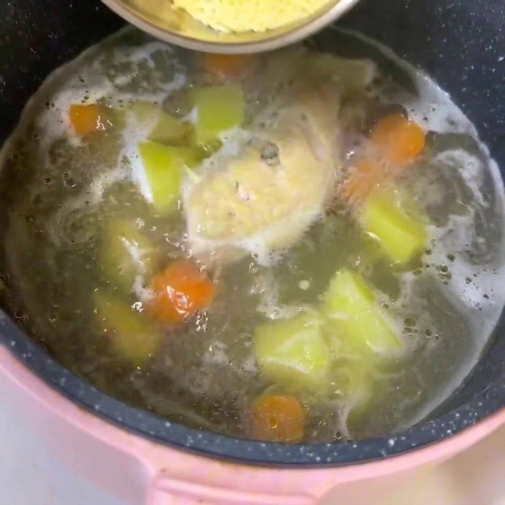 Una olla a fuego lento con caldo, pollo, patatas en dados y zanahorias, con fideos para bebés siendo incorporados.
