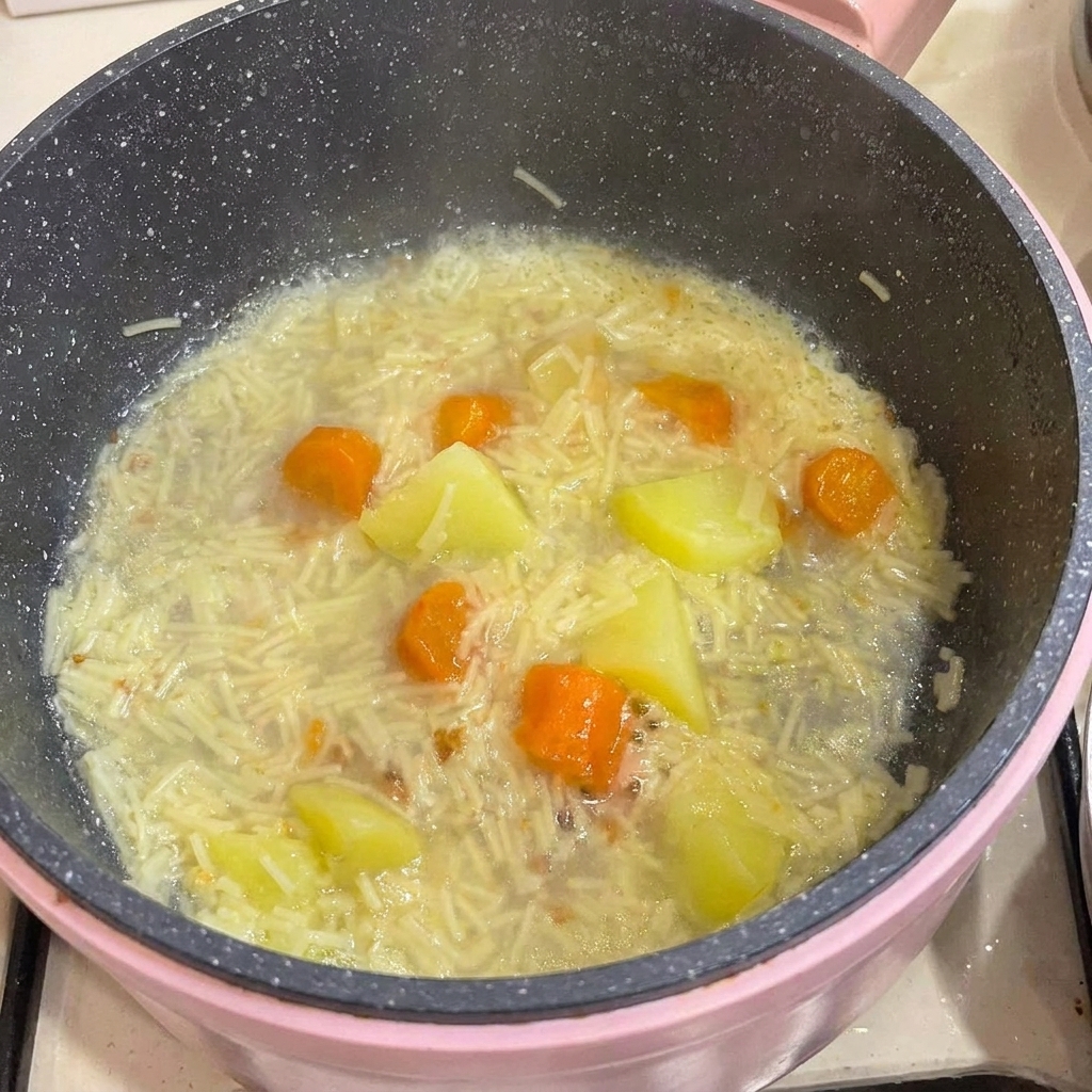 Carne de pollo deshuesada siendo añadida a una olla llena de fideos para bebés cocidos, patatas y zanahorias en caldo.