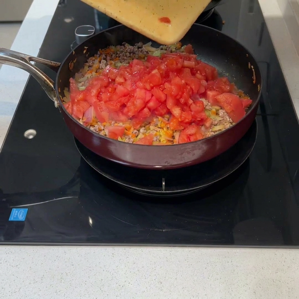 Tomates frescos picados siendo volcados desde una tabla a una sartén con carne y vegetales.