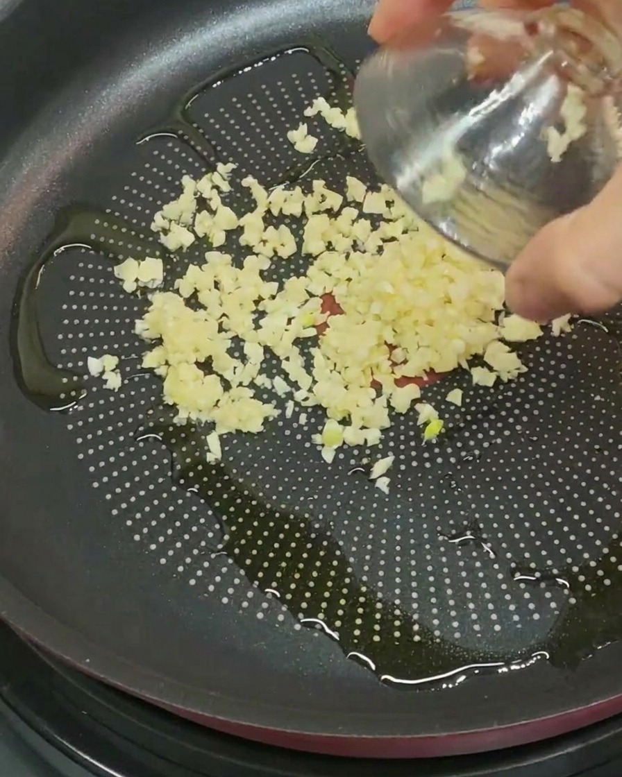 Trozos de ajo picado finamente siendo vertidos de un pequeño tazón de vidrio a una sartén con aceite de oliva.
