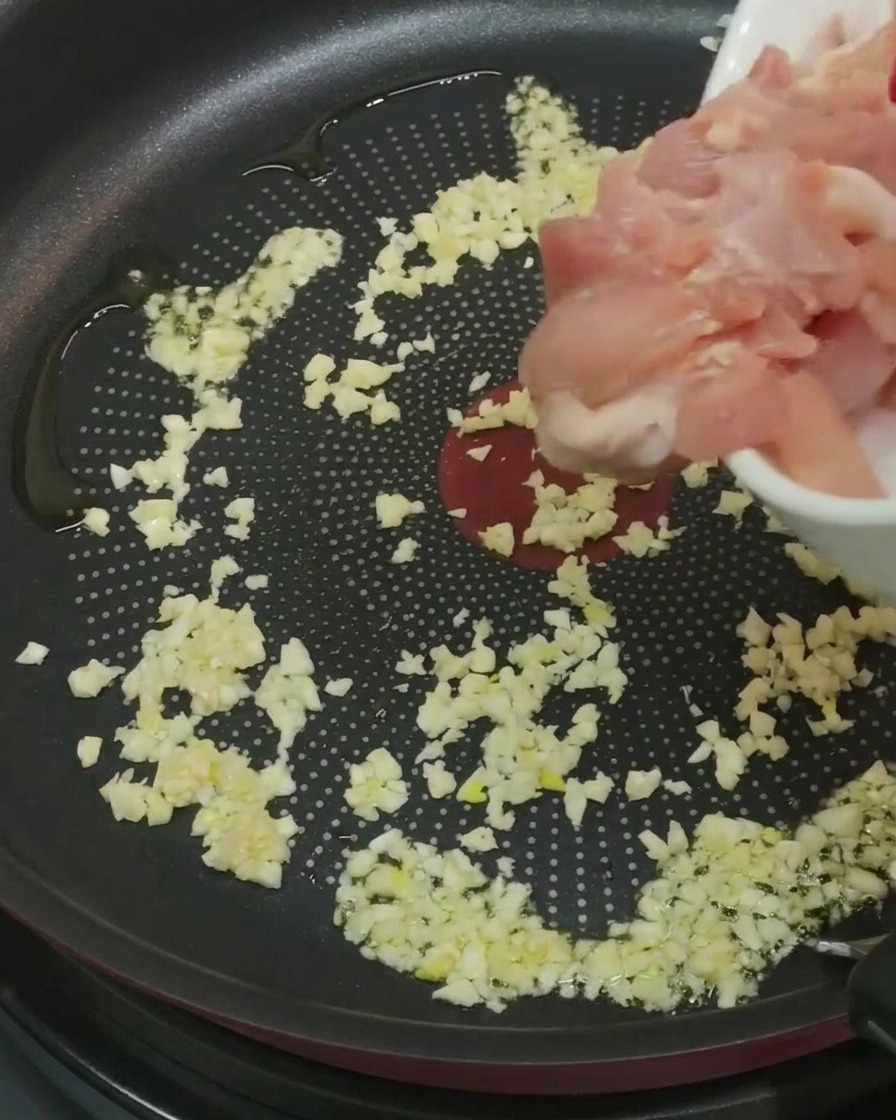 Un puñado de trozos de pollo crudo marinado siendo añadidos a una sartén que ya contiene ajo picado y aceite.