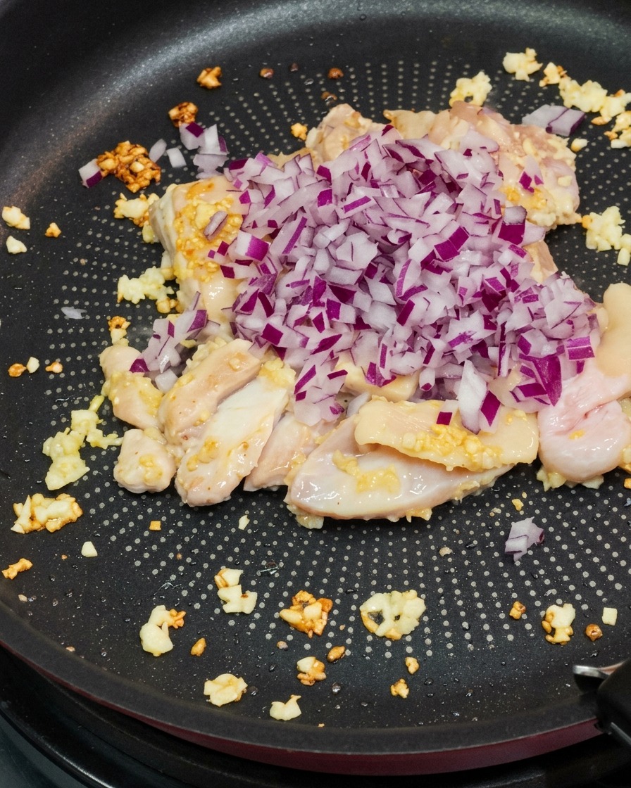 Cebollas rojas picadas siendo vertidas en una sartén con pollo salteado y ajo picado.