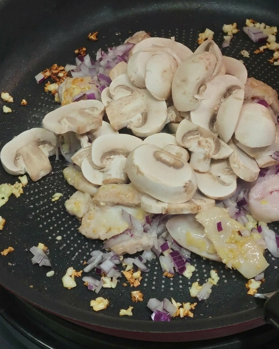 Champiñones blancos frescos en rodajas siendo añadidos a una sartén con pollo cocido, ajo y cebollas rojas.