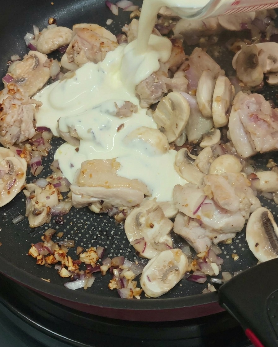Crema, leche y vino blanco siendo vertidos en la sartén con pollo cocido y champiñones y cebollas salteados.