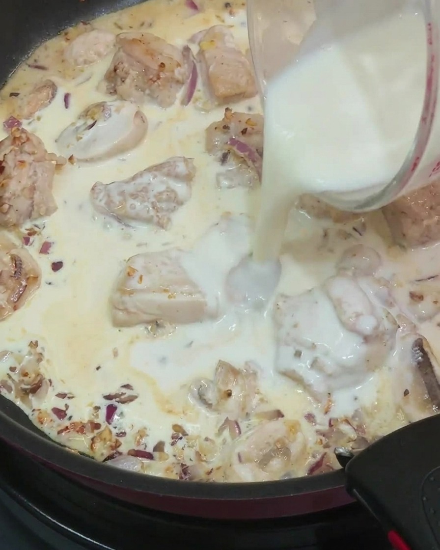 Vertiendo leche de una taza medidora de vidrio en una sartén llena con trozos de pollo cocidos, champiñones en rodajas, cebollas y crema.