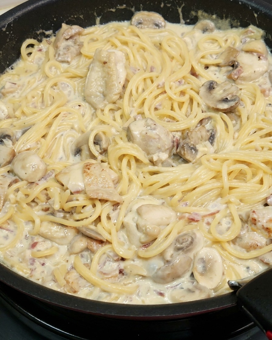 Fideos de espagueti cocidos siendo mezclados en una sartén con una rica salsa cremosa espesa, trozos de pollo y champiñones en rodajas.