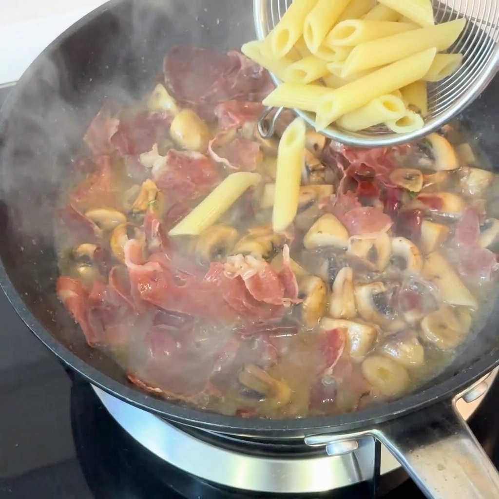 Penne cocido siendo añadido a la sartén con jamón, champiñones y salsa rica.