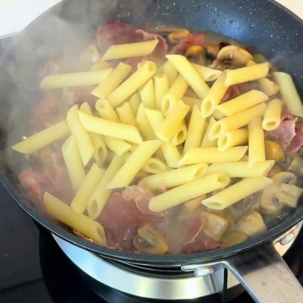 Penne cocido mezclándose en la sartén con champiñones y jamón, sazonado con pimienta negra.