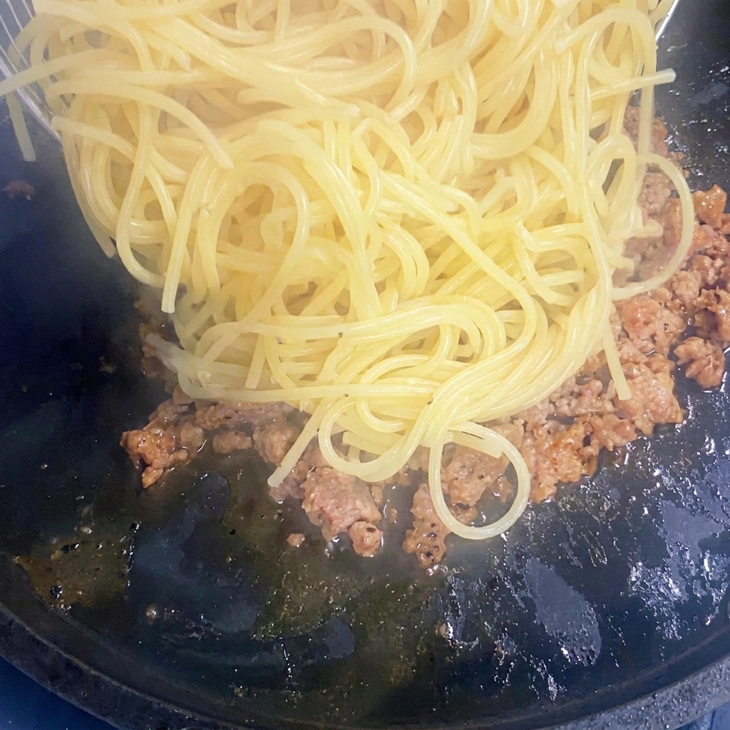 Espaguetis cocidos siendo añadidos a una sartén con carne molida dorada.