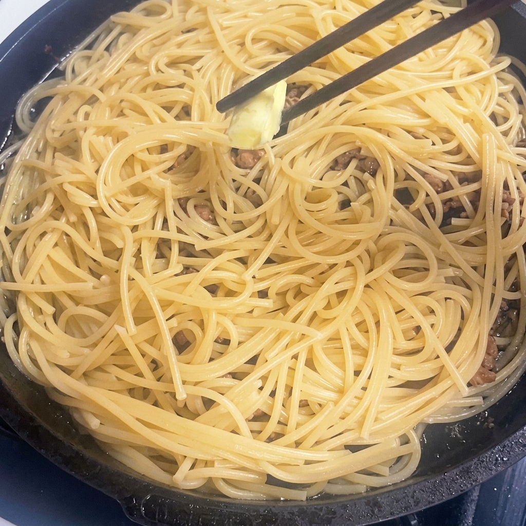 Un trozo de mantequilla derritiéndose en una sartén con pasta caliente y carne.