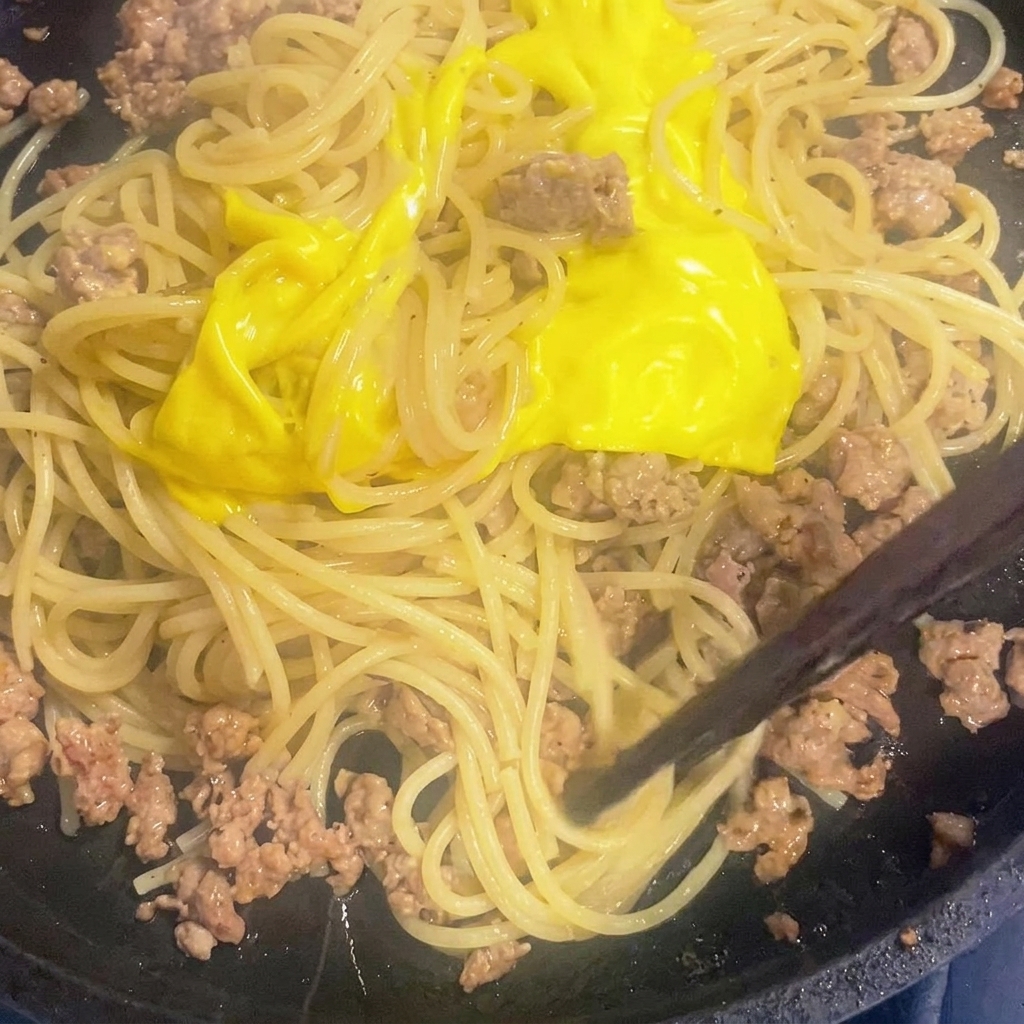 Queso derretido siendo mezclado con espaguetis y carne en una sartén.