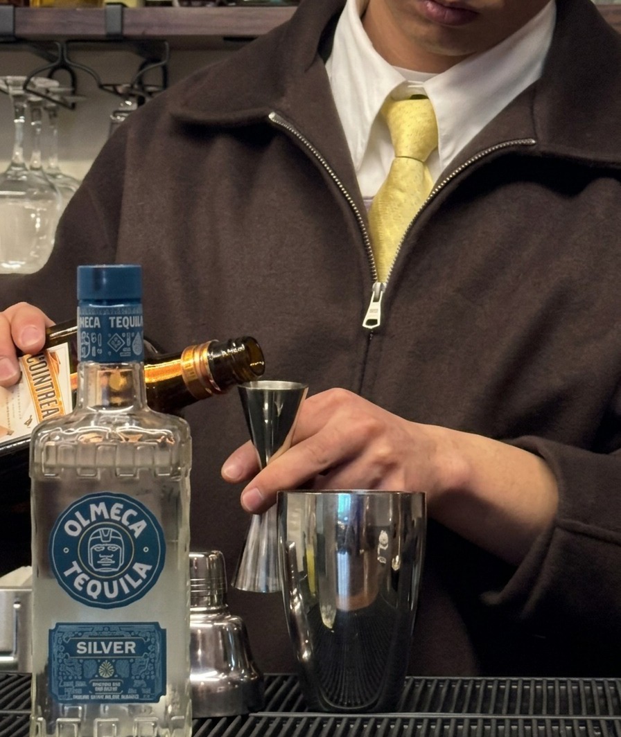 Triple sec siendo vertido de una botella a un jigger sobre una coctelera.