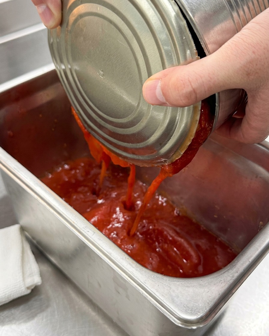 Tomates pelados en conserva siendo vertidos en una bandeja de catering de acero inoxidable limpia.