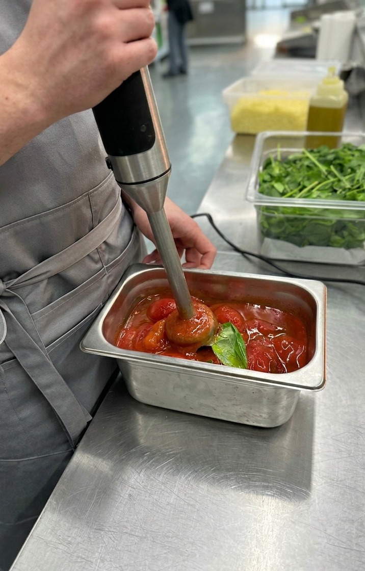 Primer plano de una batidora de mano procesando tomates pelados enteros y hojas de albahaca hasta obtener una salsa de pizza roja, vibrante y suave.