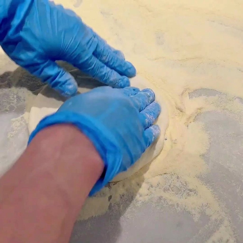 Manos con guantes azules presionando y estirando la masa cruda de pizza sobre una encimera espolvoreada con harina.