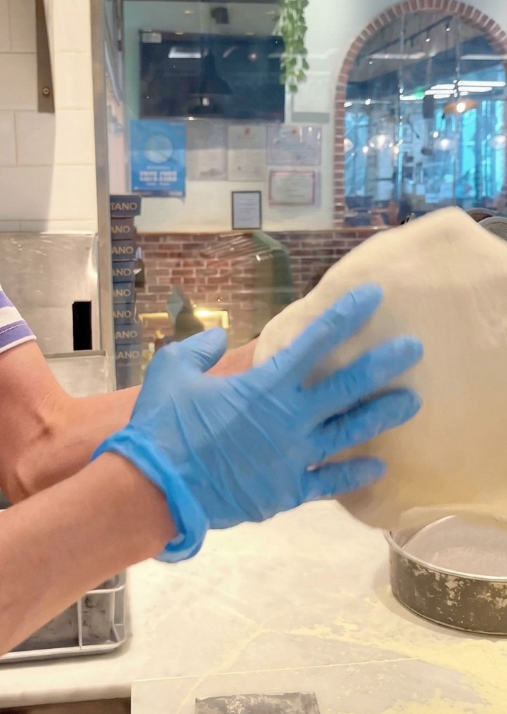 Manos con guantes azules lanzando y estirando la masa de pizza en el aire.