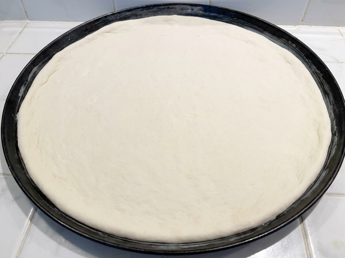 Una masa redonda de pizza cruda siendo presionada en una bandeja de hornear de metal negro.