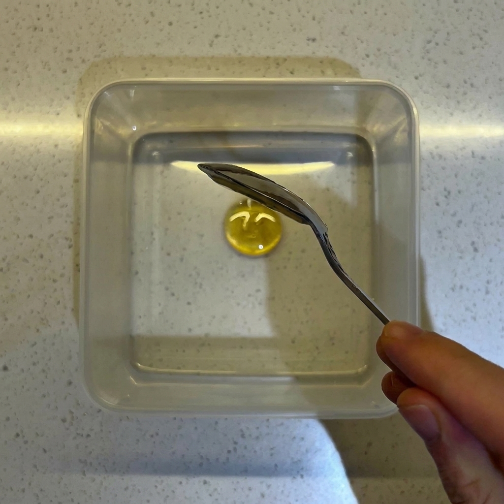 Primer plano de una cuchara mezclando miel y levadura en agua dentro de un recipiente cuadrado transparente.