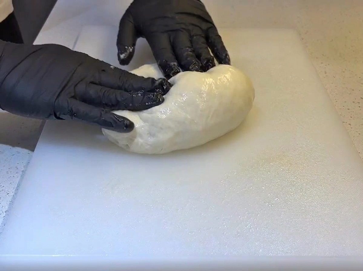 Manos con guantes negros doblando una bola de masa suave y engrasada sobre una tabla de cortar blanca.
